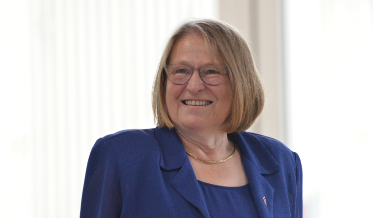 Eine Frau, dunkelblondes Haar, Brille, Kette, blaues Kostüm mit Bischofsnadel am Revers, lächelt freundlich vor sehr hellem Hintergrund.