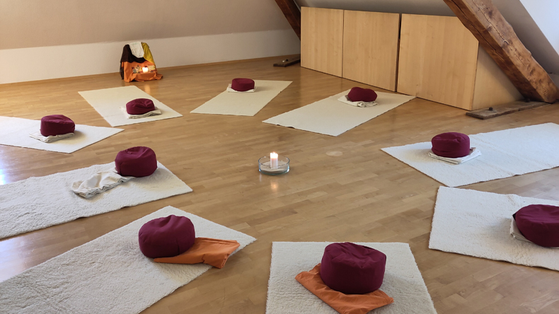 In einem Dachgeschossraum liegen rund um eine brennende Kerze neun einheitliche Teppichläufer mit jeweils einem zusammengelegten Tuch und einem runden Kissen darauf.