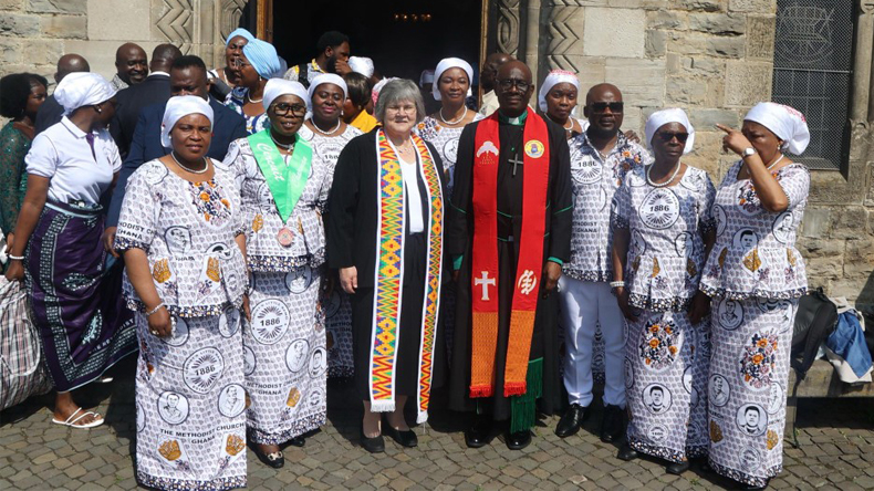Vor einem Kircheneingang stehen etwa zwei Dutzend Menschen, mehrheitlich Frauen. Einige von ihnen schauen in die Kamera. Viele von ihnen sind einheitlich gekleidet. Auf ihrer Kleidung steht der Name ihrer heimatlichen Kirche: Methodist Church Ghana.