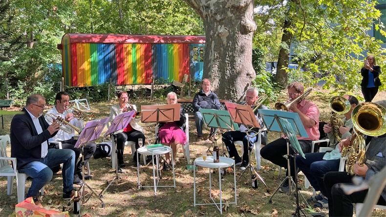 In einem Garten unter einem großen alten Laubbaum spielt ein achtköpfiger Posaunenchor: zwei Frauen und sechs Männer. Im Hintergrund steht ein bunt bemalter Wagen.