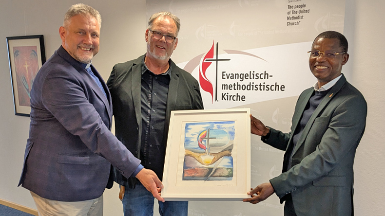 Im Flur der Kirchenkanzlei halten drei Herren in Jacketts gemeinsam ein gerahmtes Gemälde. Es zeigt Kreuz und Flamme sowie das Kreuz in der Weltkugel über zwei offenen Händen, durch die helles Licht scheint. Die unten dargestellte Welt ist zerrissen.