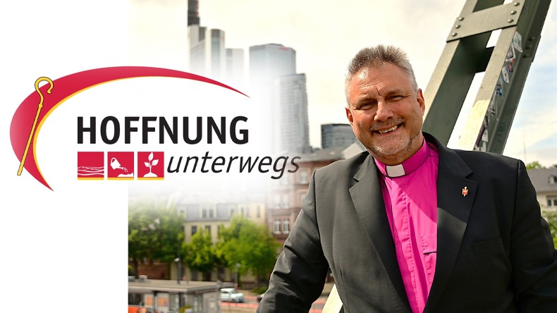 Ein lächelnder Geistlicher in dunklem Sakko und violettem Kollarhemd steht auf einer Brücke vor moderner Stadtkulisse. Links im Bild erscheint das Logo der Initiative »Hoffnung unterwegs«.