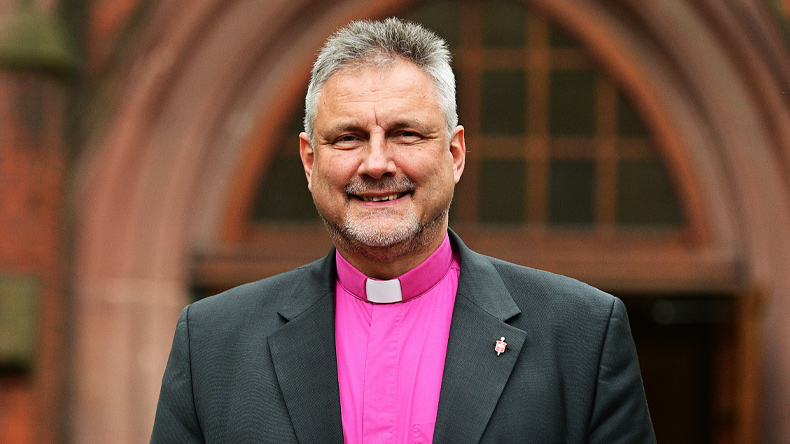 Werner Philipp steht vor dem Eingang der Christuskirche am Merianplatz in Frankfurt am Main. Er lächelt freundlich und direkt in die Kamera: kurze graumelierte Haare und ebensolcher Bart, Kollarhemd und dunkles Jackett mit Bischofsnadel im Revers.