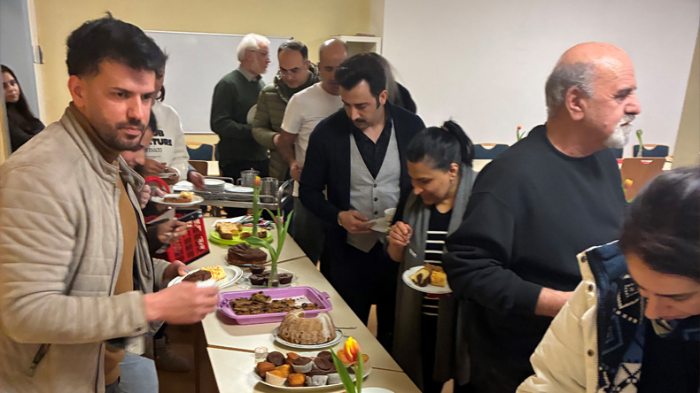 Beiderseits eines langen Tisches stehen Menschen unterschiedlichen Alters und Geschlechts. Sie laden sich Kuchen und anderes süßes Gebäck auf ihre Teller. Der Tisch ist mit Tulpen geschmückt.