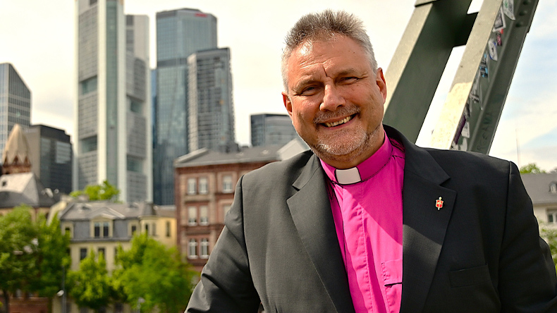 Bischof Philipp steht auf einer Mainbrücke in Frankfurt. In Kollarhemd und dunklem Jackett lächelt er freundlich in die Kamera. Im Hintergrund einige Wolkenkratzer von Banken und einige Stadthäuser sowie belaubte Bäume.