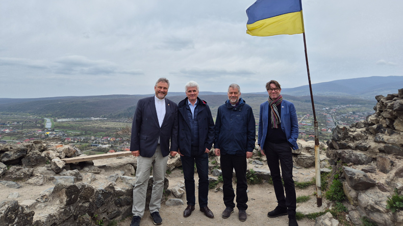 	Vier Männer stehen auf einem Berg und lächeln in die Kamera. Rechts neben ihnen weht eine ukrainische Flagge: oben blau, unten gelb. Im Tal stehen viele niedrige Häuser und fließt ein Fluss.