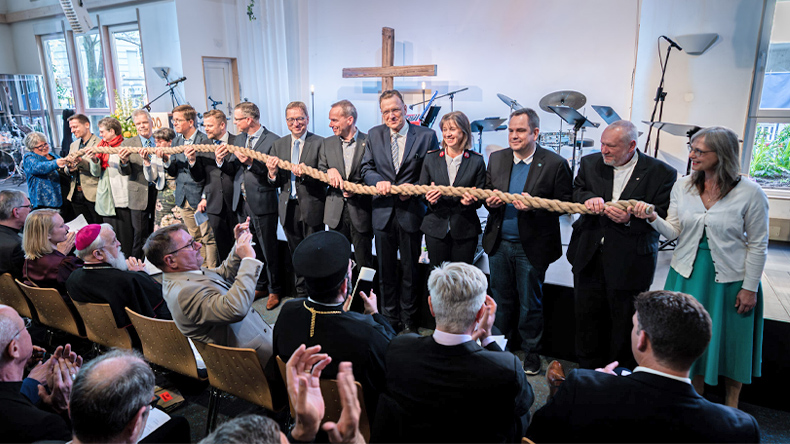 Fünfzehn Menschen halten gemeinsam ein Tau. Sie stehen in einem Gottesdienstraum vorne und den Festgottesdienstbesuchern zugewandt. Hinter ihnen steht ein Kreuz.