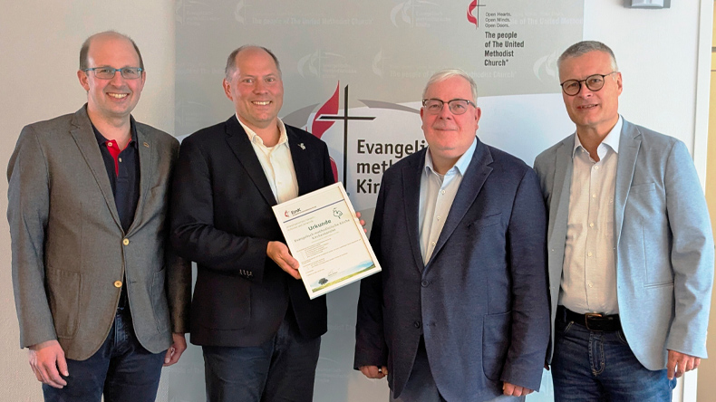 Vier Männer stehen vor einem stehenden Banner der Evangelisch-methodistischen Kirche. Einer von ihnen hält die Urkunde zur erneuten Auszeichnung der Kirchenkanzlei mit dem Grünen Gockel in der Hand.