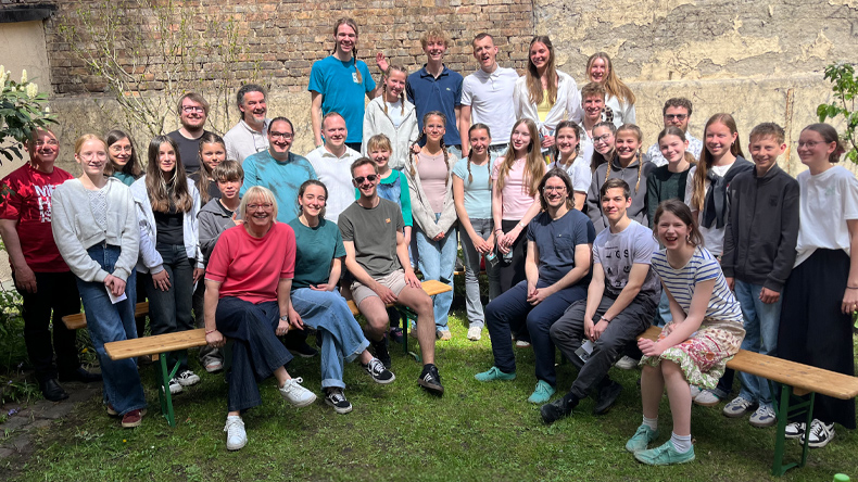 Etwas über dreißig meist junge Menschen haben sich zu einem Gruppenfoto in einem begrünten Hinterhof versammelt und lächeln freundlich in die Kamera. Im Hintergrund ist eine Ziegelmauer.
