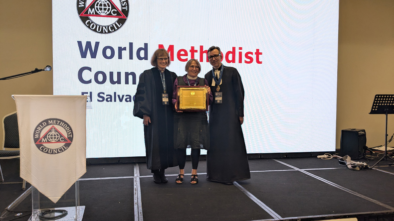 Drei Personen stehen auf einer Bühne vor einer großen Leinwand mit der Aufschrift 'World Methodist Council El Salvador'. Zwei Personen tragen lange, dunkle Talare, die mittlere Person (Rosemarie Wenner) hält eine gerahmte Urkunde und trägt Alltagskleidung. Alle drei lächeln. Links steht ein Rednerpult mit einem weißen Tuch, auf dem das Logo des World Methodist Council zu sehen ist. Im Hintergrund rechts steht ein Notenständer mit Mikrofon.