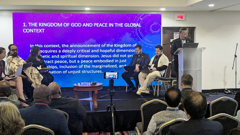 Teilnehmende sitzen vor einer Bühne, auf der sechs Personen über Frieden im weltweiten Zusammenhang sprechen. Im Hintergrund ist eine englischsprachige Präsentation zum Reich Gottes und zum Frieden zu sehen.